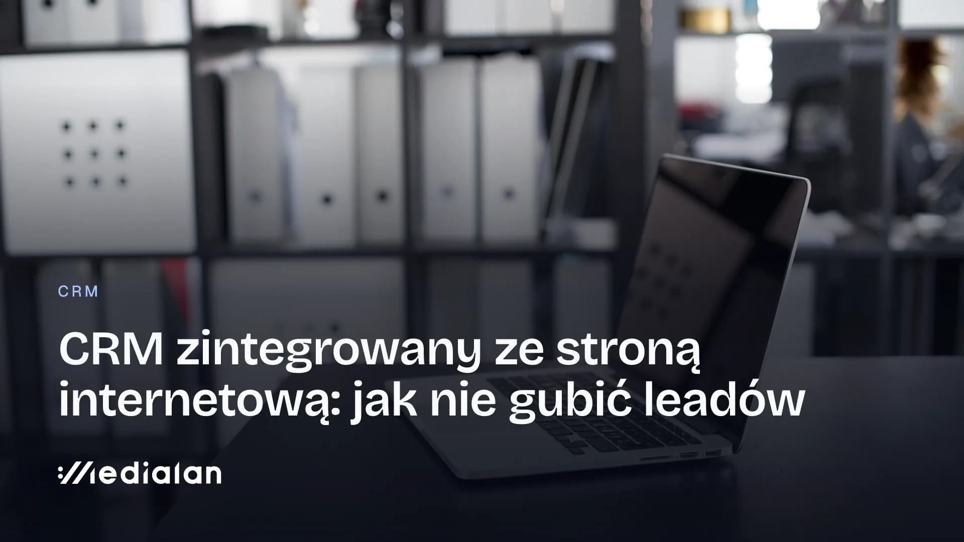Otwarty laptop na ciemnym biurku w biurze, w tle regał z segregatorami i pudełkami na dokumenty